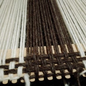 May include: A weaving project on a rigid heddle loom featuring vertical warp threads in alternating stripes of cream, dark brown, and light gray. A horizontal dark brown weft thread is woven through the warp to create a plain weave pattern.