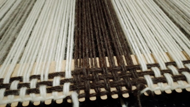 May include: A weaving project on a rigid heddle loom featuring vertical warp threads in alternating stripes of cream, dark brown, and light gray. A horizontal dark brown weft thread is woven through the warp to create a plain weave pattern.