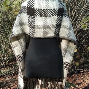 May include: A woven shawl featuring a thick, textured plaid pattern in shades of cream, dark brown, and grey. The garment has a wide, draped silhouette with long, loose fringe detailing along the bottom edge, showcasing a heavy, chunky knit construction.