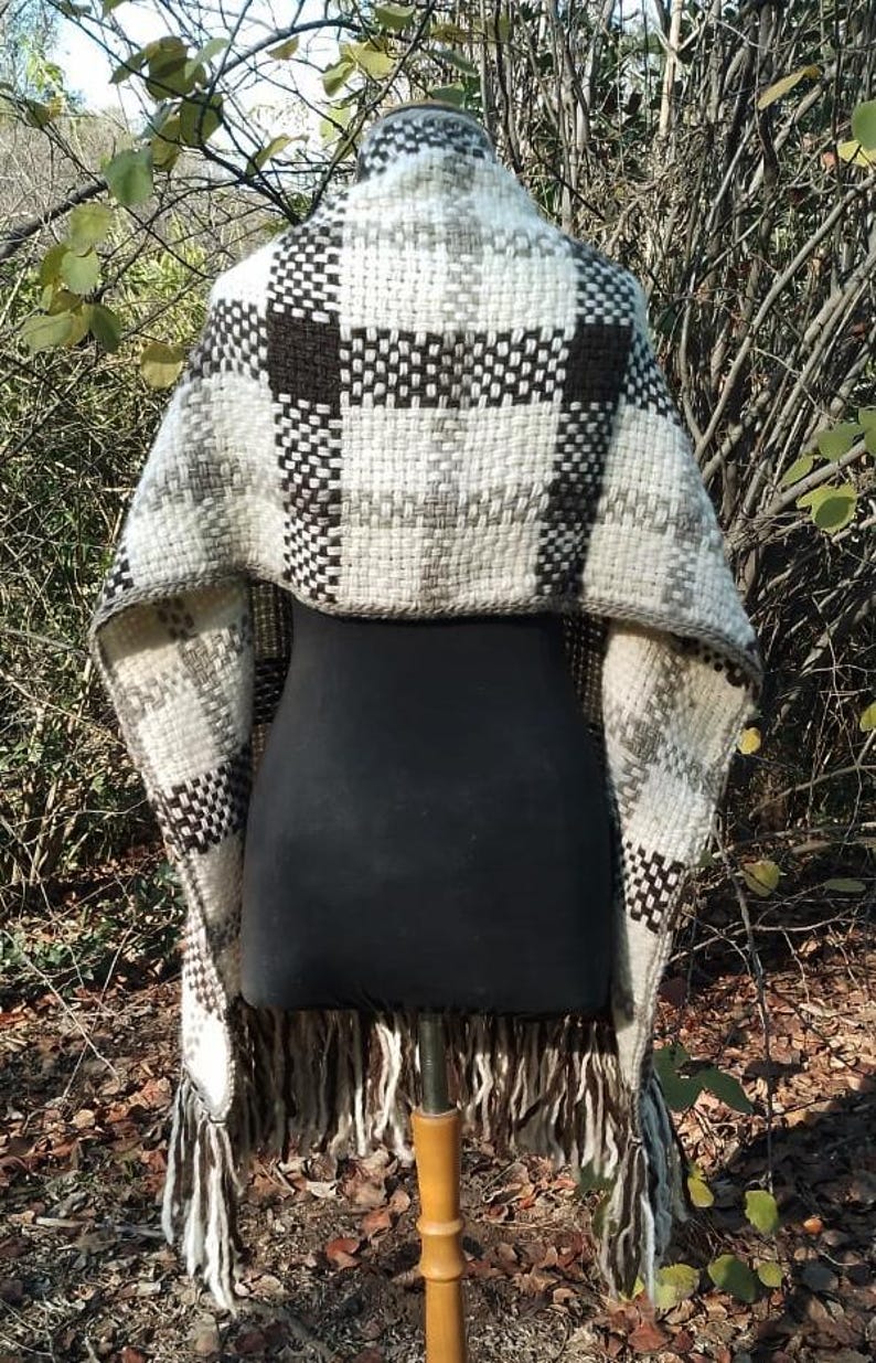 May include: A woven shawl featuring a thick, textured plaid pattern in shades of cream, dark brown, and grey. The garment has a wide, draped silhouette with long, loose fringe detailing along the bottom edge, showcasing a heavy, chunky knit construction.