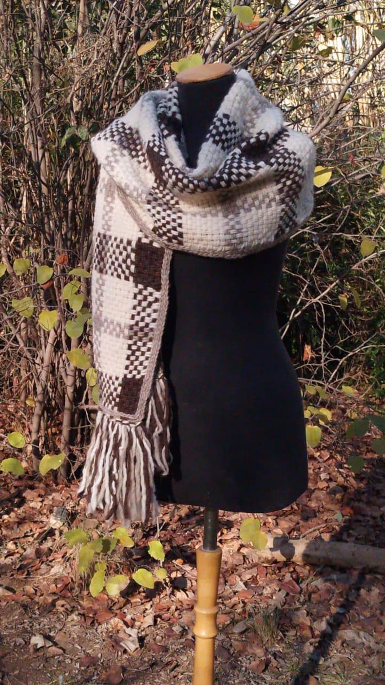 May include: A thick, woven scarf featuring a checkered pattern in shades of cream, beige, and dark brown. The scarf has a textured, chunky knit appearance with long, loose fringe detailing at the ends, draped over a black display mannequin.