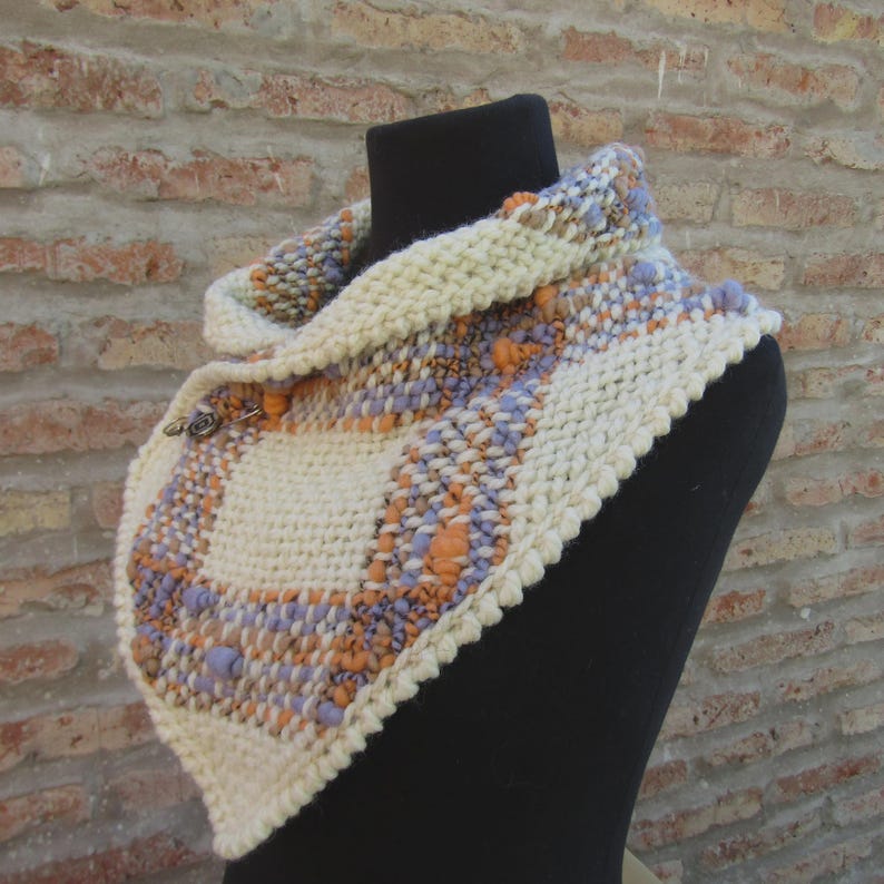 May include: A chunky knit cowl scarf featuring a cream-colored base with woven accents in lavender and orange. The scarf has a textured, multi-colored pattern and is secured at the front with a small, circular metal button fastener.