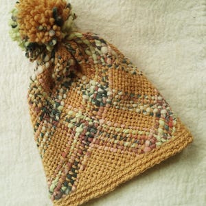 May include: A knitted beanie with a pom-pom on top, featuring a textured weave in shades of mustard yellow, sage green, soft pink, and dark grey. The hat has a solid mustard yellow ribbed brim and a multi-colored yarn pattern throughout the body.