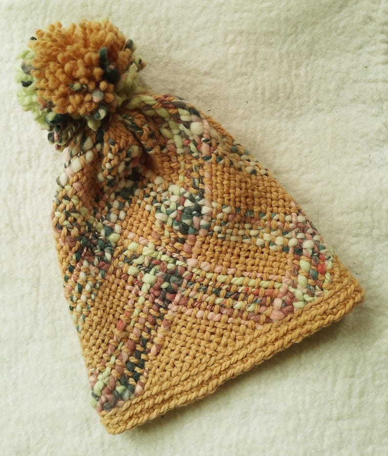 May include: A knitted beanie with a pom-pom on top, featuring a textured weave in shades of mustard yellow, sage green, soft pink, and dark grey. The hat has a solid mustard yellow ribbed brim and a multi-colored yarn pattern throughout the body.