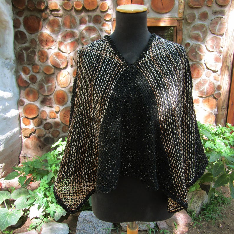 May include: A black and tan woven poncho featuring a V-neckline and a textured, grid-like pattern. The garment displays a solid black center panel that transitions into a contrasting black and tan woven design on the sides, creating a layered look.