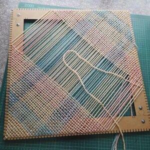 May include: A square weaving loom frame made of light brown material with notched edges, featuring a partially completed woven pattern of pastel pink, blue, and yellow yarn threads arranged in a diagonal grid across the open center space.