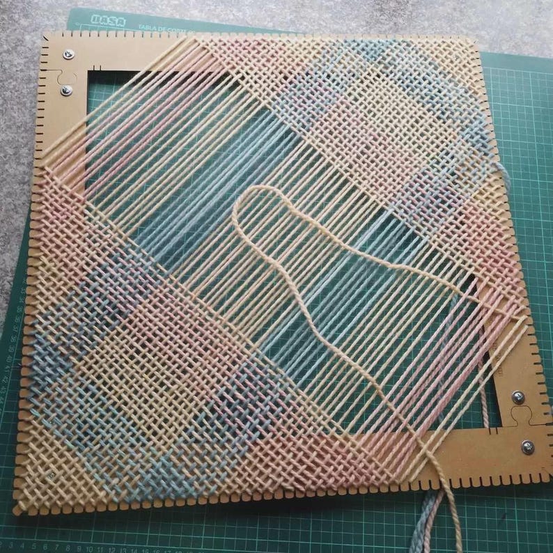 May include: A square weaving loom frame made of light brown material with notched edges, featuring a partially completed woven pattern of pastel pink, blue, and yellow yarn threads arranged in a diagonal grid across the open center space.