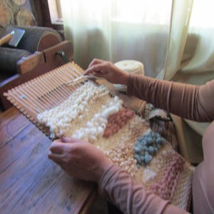 May include: A wooden frame loom with a partially woven textile featuring thick, textured yarn in shades of cream, dusty pink, and muted blue. The weaving displays varied loop and knot patterns, with a ball of cream yarn resting nearby.