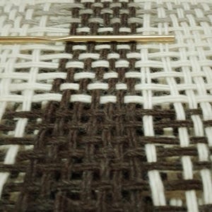 May include: A woven textile project featuring a geometric pattern of dark brown, white, and light gray yarn threads. A thin, metallic gold-colored crochet hook is positioned horizontally across the center of the woven fabric structure.