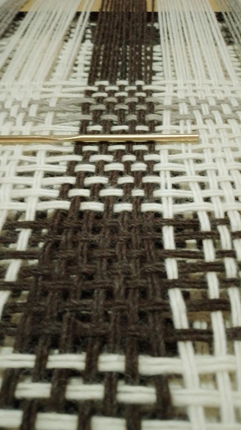 May include: A woven textile project featuring a geometric pattern of dark brown, white, and light gray yarn threads. A thin, metallic gold-colored crochet hook is positioned horizontally across the center of the woven fabric structure.