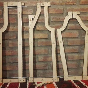 May include: Three light-colored wooden weaving loom frames with notched edges and metal rods, arranged vertically. Each frame features printed text and markings. A small wooden comb tool is positioned horizontally at the bottom center.