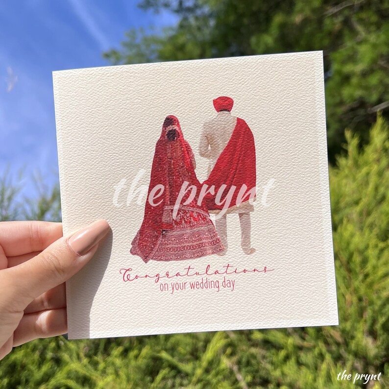 Puede incluir: Tarjeta de boda cuadrada con una ilustraci&oacute;n de una pareja con vestimenta tradicional. La novia lleva un vestido y velo rojos, y el novio un atuendo crema con un chal rojo. El texto "Felicidades en tu d&iacute;a de boda" est&aacute; escrito en la tarjeta.