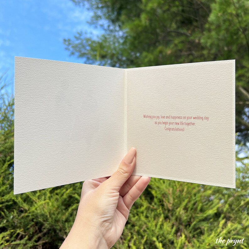 Puede incluir: Tarjeta de boda abierta con una superficie texturizada de color blanquecino. El interior presenta texto rojo: &laquo;Dese&aacute;ndoles alegr&iacute;a, amor y felicidad en el d&iacute;a de su boda, al comenzar su nueva vida juntos. &iexcl;Felicidades!&raquo;. La tarjeta se sostiene contra follaje verde.