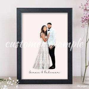 May include: A framed illustration of a couple in wedding attire. The bride is wearing a white gown with a floral pattern and the groom is wearing a white tuxedo jacket with black pants. The couple is standing close together and the groom has his arm around the bride. The illustration is on a pink background and the couple's names, "Giran & Dalveer", are written in cursive at the bottom.