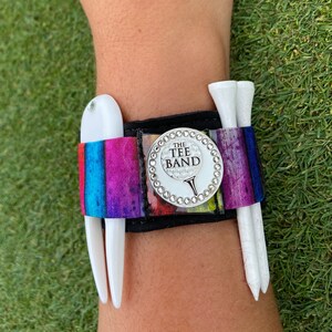 May include: A black and white striped golf wristband with a silver charm that says "The Tee Band". The wristband has two white golf tees attached to it.