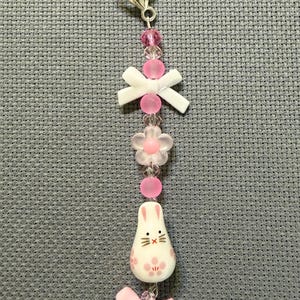 May include: A handmade keychain featuring a white bunny charm with pink floral accents and a pink bow. The keychain also includes pink and clear beads, a white bow, and a silver-tone clasp.