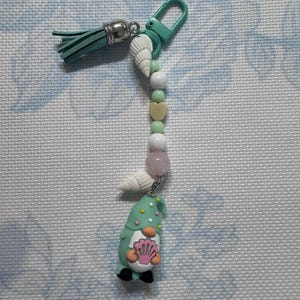 May include: A teal keychain with a gnome charm holding a pink seashell. The keychain features a teal clasp, a tassel, and a strand of beads including white, green, and pink beads, and seashells. The gnome is wearing a teal hat with white dots.