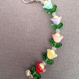 May include: A beaded charm with a silver clasp, featuring colourful flower beads in shades of blue, purple, pink, yellow, and red. Each flower has green leaf accents and is strung with clear beads. The charm is on a grey background.