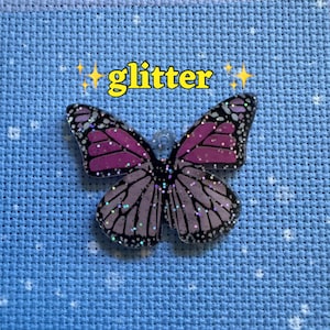 May include: A glittery butterfly-shaped charm with pink and black wings. The word "glitter" is written in yellow above the butterfly. The charm is set against a light blue background.