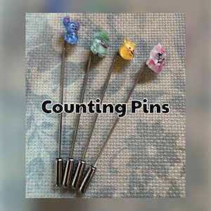 May include: Four counting pins with different cartoon character toppers. The toppers are blue, green, yellow, and pink. The pins are silver and have a rounded end.