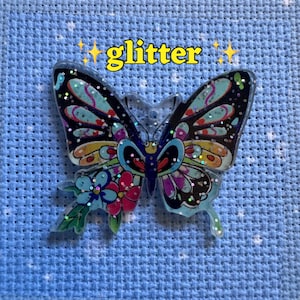 May include: A decorative butterfly sticker with glitter accents. The butterfly features a vibrant design with black, teal, pink, and yellow wings. The word "glitter" is written in yellow above the butterfly.