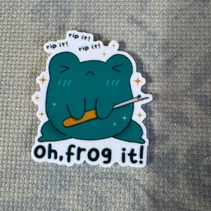 May include: A green frog cartoon sticker with the text "rip it!" repeated three times and the text "oh.frog it!" at the bottom. The frog is holding a needle and thread.