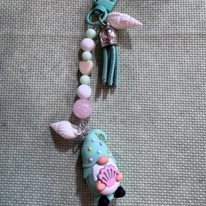 May include: A whimsical keychain featuring a turquoise gnome with a pink seashell, adorned with a beaded strand of pink and green beads, a turquoise tassel, and a seashell charm. The keychain has a turquoise clasp.
