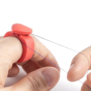 May include: A red plastic ring with a small, heart-shaped top that holds thread. The ring is worn on a finger and helps to keep the thread taut while sewing.