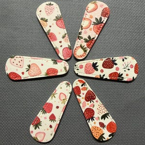May include: Six white and pink hair clips with a strawberry pattern. The clips are in various shapes, with a repeating design of red strawberries, green leaves, and small white flowers. The clips are arranged in a circular pattern.