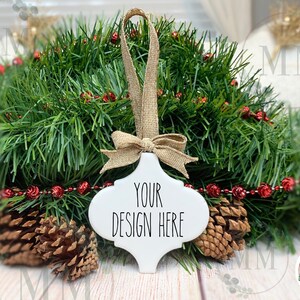 May include: White ceramic Christmas ornament with a rustic twine bow and the text "Your Design Here". The ornament is surrounded by a green garland with red berries and pine cones.