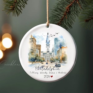 May include: A round, white ceramic Christmas ornament featuring a watercolor painting of Philadelphia's cityscape. The ornament includes the city name, names, and the year 2024 with a heart symbol. A brown string is attached for hanging.
