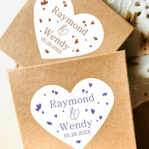 May include: Two square brown cardboard boxes, each featuring a white heart-shaped sticker. The top sticker has brown text reading Raymond & Wendy 05.28.20XX with brown hearts. The bottom sticker has purple text and purple hearts.