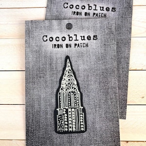 May include: Two iron-on patches featuring a black and white illustration of a skyscraper. The patches are on a gray denim-textured background. The text "Cocoblues IRON ON PATCH" is printed above each patch.