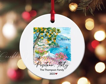 May include: White ceramic ornament with a watercolor painting of Positano, Italy. The artwork features a coastal scene with a tree bearing yellow fruit, buildings, and the text "Positano, Italy". Includes "The Thompson Family 2025".