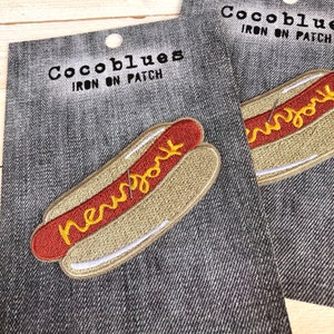 May include: Embroidered iron-on patch featuring a hot dog with the word "New York" in yellow script. The patch has a red hot dog, yellow mustard, and a tan bun. The patch is on a denim background.