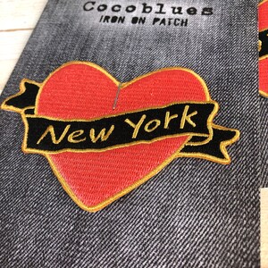 May include: An embroidered patch with a red heart and a black banner displaying "New York" in gold lettering. The patch has a gold border and is set against a denim background. The text "Cocoblues IRON ON PATCH" is visible at the top.