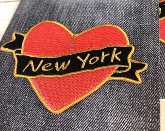 Peut inclure: Un écusson brodé avec un cœur rouge et une bannière noire avec l'inscription "New York" en lettres dorées. L'écusson a une bordure dorée et est posé sur un fond en denim. Le texte "Cocoblues IRON ON PATCH" est visible en haut.