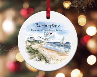 Könnte beinhalten: Runder, weißer Keramik-Weihnachtsschmuck mit Küstenmotiv. Der Schmuck zeigt einen Strand mit Segelboot, Häusern und dem Text "The Hamptons" und Namen. Das Jahr 2024 ist ebenfalls enthalten, zusammen mit einem Herzsymbol.