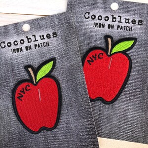 以下が含まれることがあります： 赤いリンゴのデザイン、黒いアウトライン、緑の葉、そして「NYC」の文字が特徴の2つのアイロン接着パッチ。デニム調の背景に配置され、上部には「Cocoblues IRON ON PATCH」の文字があります。