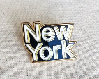 May include: A rectangular pin with the words "New York" in white, outlined in gold, set against a black background. The pin has a gold border and is likely a souvenir or collectible item.