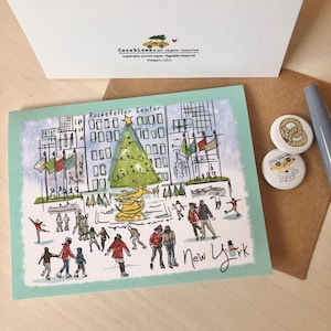 Può includere: Un biglietto d'auguri a tema natalizio con un'illustrazione ad acquerello del Rockefeller Center a New York City, con un albero di Natale e pattinatori. Il biglietto è accompagnato da due spille rotonde e una penna. Il biglietto dice "New York".