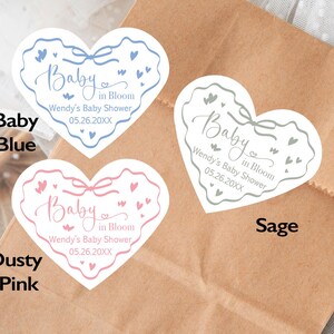 May include: Heart-shaped stickers in baby blue, dusty pink, and sage green. Each sticker features the text "Baby in Bloom" and "Wendy's Baby Shower 05.26.20XX". The stickers have a bow and heart design.