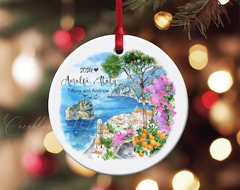 May include: A round white ceramic Christmas ornament with a watercolor painting of Amalfi, Italy. The ornament has a red ribbon for hanging and features the text "2024 Amalfi, Italy" and the names "Tiffany and Andrew."