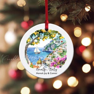 Pode incluir: Enfeite de Natal redondo em cerâmica branca com uma pintura em aquarela de Amalfi, Itália. A imagem inclui um veleiro, edifícios costeiros e o texto "Amalfi, Italy" e "2026". Uma fita vermelha é anexada para pendurar.