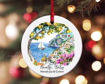 Op de afbeelding: Ronde witte keramische kerstversiering met een aquarel schilderij van Amalfi, Italië. De afbeelding toont een zeilboot, kustgebouwen en de tekst "Amalfi, Italy" en "2026". Een rood lint is bevestigd om op te hangen.