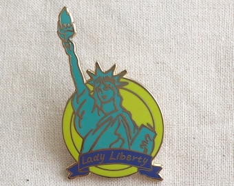 May include: A teal and gold enamel pin featuring the Statue of Liberty. The pin has a yellow background and a blue banner with the words "Lady Liberty". The torch is lit with a flame.