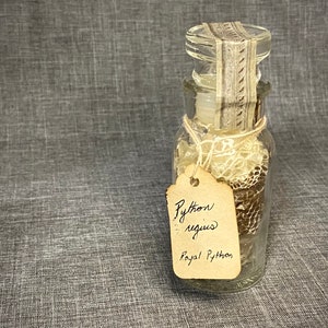 May include: A clear glass jar containing snake skin and a tag that reads "Python regius Royal Python". The jar is sealed with a decorative label and tied with twine. The snake skin has brown and tan patterns.