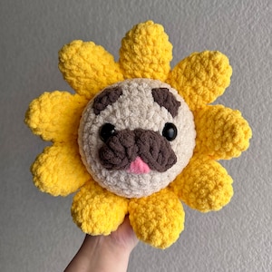 May include: A crocheted plush toy shaped like a sunflower with a pug face in the center. The sunflower is yellow and the pug is brown and white.