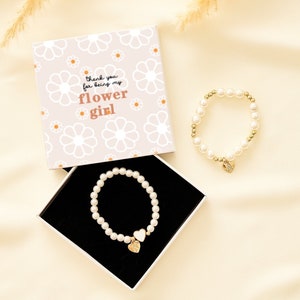 May include: A white box with a floral pattern and the text "Thank you for being my flower girl" and a white pearl bracelet with a gold heart charm in a black box.