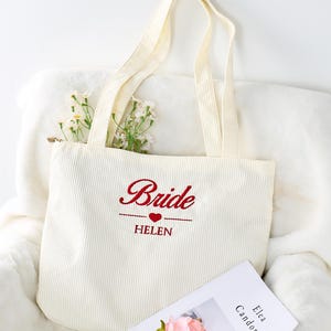 Può includere: Borsa tote in velluto a coste color crema con la scritta "Bride" ricamata in rosso, un cuore rosso e il nome "Helen". La borsa ha due manici ed è piena di fiori bianchi. Una rivista e un fiore rosa sono in primo piano.